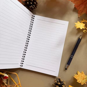 May include: A lined notebook with a spiral binding, a black pen, pine cones, and autumn leaves. The notebook is on a brown surface, surrounded by fall foliage in shades of orange, yellow, and brown.