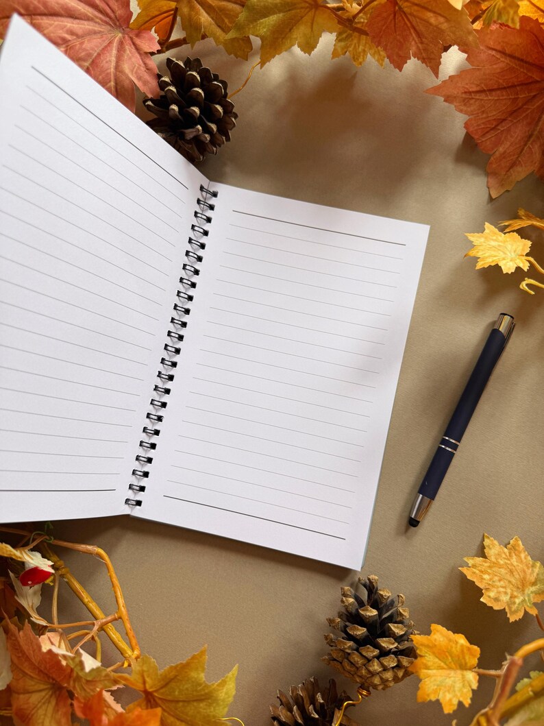 May include: A lined notebook with a spiral binding, a black pen, pine cones, and autumn leaves. The notebook is on a brown surface, surrounded by fall foliage in shades of orange, yellow, and brown.