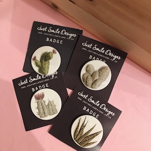 May include: Four round badges with black card stock backing. Each badge features a different cactus or succulent illustration. The text "Just Smile Designs" and "Badge" are printed on each card.