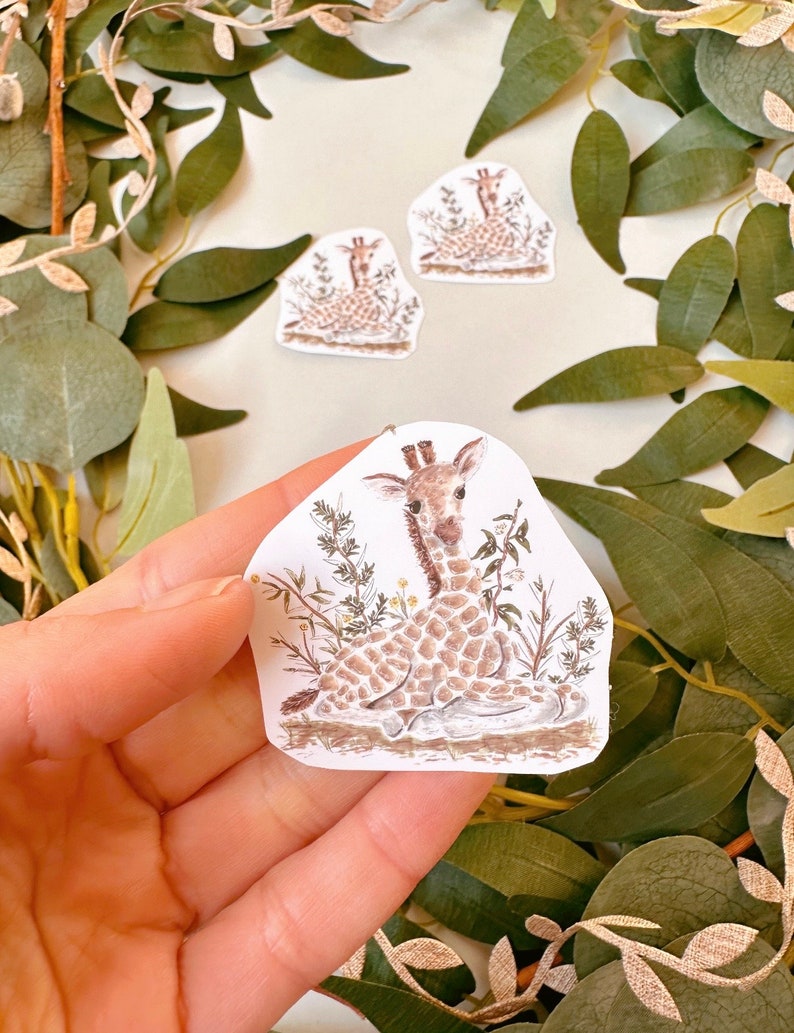 May include: A sticker featuring a hand-drawn illustration of a baby giraffe sitting in a patch of green grass. The giraffe is brown and white with a long neck and large ears. The sticker is on a white background.