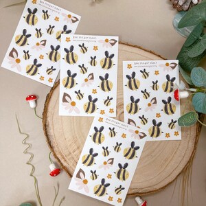 May include: Four sheets of bee stickers with a yellow and black bee design. The stickers are on a white background with a brown wood surface behind them. The text on the stickers reads "Bee Sticker Sheet" and "www.justminimaldesigns.co.uk".