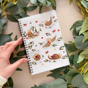 May include: A white spiral bound notebook with a colorful illustration of sea otters, seaweed, and red crabs. The otters are brown and white and are depicted in various poses, including swimming, holding hands, and playing with seaweed.
