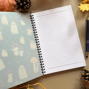 May include: Open spiral notebook with lined pages and a patterned cover featuring white ghosts and mushrooms on a light blue background. A black pen rests on the right side. Autumn leaves and pinecones surround the notebook.