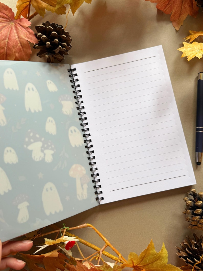 May include: Open spiral notebook with lined pages and a patterned cover featuring white ghosts and mushrooms on a light blue background. A black pen rests on the right side. Autumn leaves and pinecones surround the notebook.