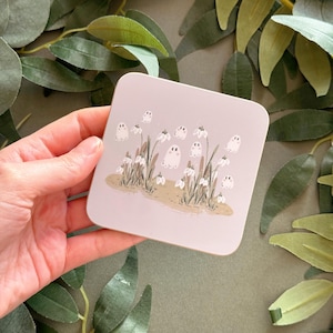 May include: A square coaster with a light purple background featuring a whimsical illustration of white snowdrops and small white ghosts. The snowdrops are clustered together in a field with brown stems and green leaves. The ghosts are hovering above the flowers, some with small, black eyes.