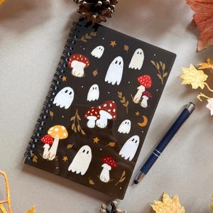 May include: A spiral-bound notebook with a dark brown cover featuring white ghost and red mushroom illustrations. A blue pen rests beside the notebook. Autumn leaves and pinecones surround the notebook, creating a seasonal theme.