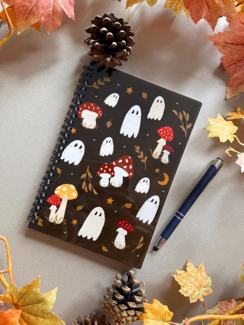 May include: A spiral-bound notebook with a dark brown cover featuring white ghost and red mushroom illustrations. A blue pen rests beside the notebook. Autumn leaves and pinecones surround the notebook, creating a seasonal theme.