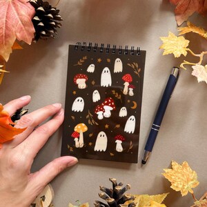 May include: A dark brown spiral-bound notebook with illustrations of white ghosts and red and yellow mushrooms. A dark blue pen lies beside the notebook. The scene is decorated with autumn leaves and a pinecone. The notebook is approximately 10 cm x 15 cm.