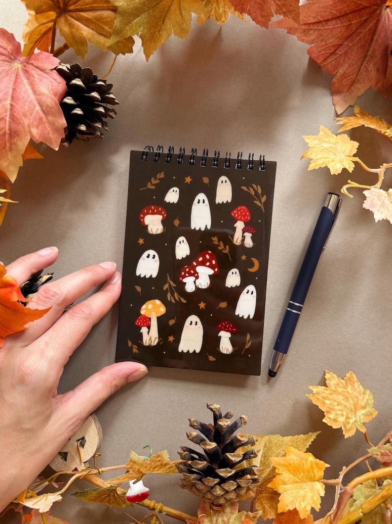 May include: A dark brown spiral-bound notebook with illustrations of white ghosts and red and yellow mushrooms. A dark blue pen lies beside the notebook. The scene is decorated with autumn leaves and a pinecone. The notebook is approximately 10 cm x 15 cm.