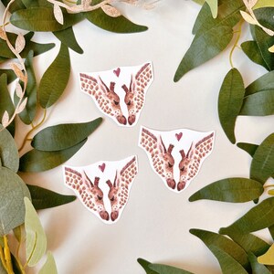 May include: Three watercolor illustrations of two giraffes kissing with a small red heart above them. The giraffes are brown and white with a light brown background.
