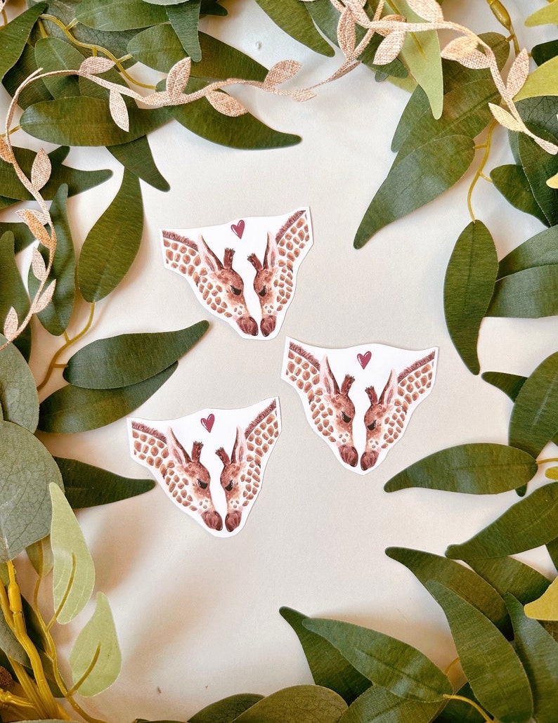 May include: Three watercolor illustrations of two giraffes kissing with a small red heart above them. The giraffes are brown and white with a light brown background.
