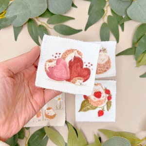 Puede incluir: Cuatro cuadrados de tela blanca con im&aacute;genes bordadas de erizos. Un cuadrado presenta dos erizos con corazones rojos. Otro cuadrado muestra un erizo con fresas. Los dos cuadrados restantes muestran erizos con flores.