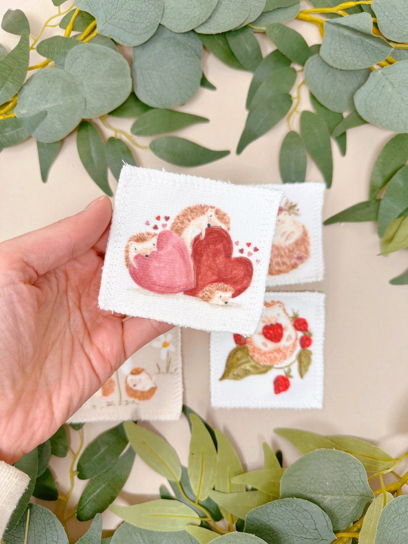 Puede incluir: Cuatro cuadrados de tela blanca con im&aacute;genes bordadas de erizos. Un cuadrado presenta dos erizos con corazones rojos. Otro cuadrado muestra un erizo con fresas. Los dos cuadrados restantes muestran erizos con flores.