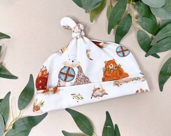 May include: A white baby hat with a knotted top, featuring a repeating pattern of woodland animals, including bears and hedgehogs. The illustrations are in shades of brown, orange, and blue, set against a white background. The hat is surrounded by green eucalyptus leaves.