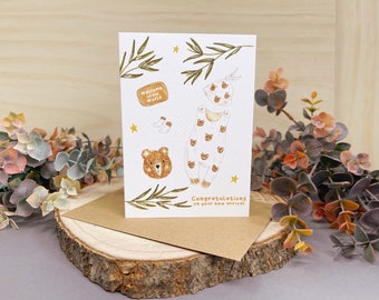 Op de afbeelding: Een witte kaart met een bruine envelop. De kaart heeft een handgetekende illustratie van een baby-outfit met een berenpatroon. De tekst op de kaart luidt "Welkom op de wereld" en "Gefeliciteerd met jullie nieuwe aanwinst".