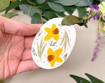 May include: A white ceramic oval ornament with a green ribbon. The ornament is painted with two yellow daffodils with brown centers and green leaves.