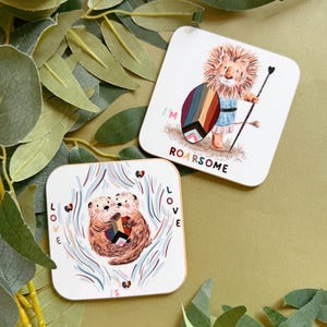 May include: Two square coasters with animal illustrations. One features a lion with a shield and spear, the text reads "I'M ROARSOME". The other shows two otters with the text "LOVE IS LOVE". Both have a white background.