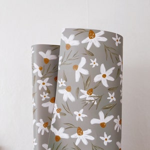 Op de afbeelding: Een rol grijs inpakpapier met een wit madeliefje bloemenpatroon. De madeliefjes hebben gele centra en groene bladeren.