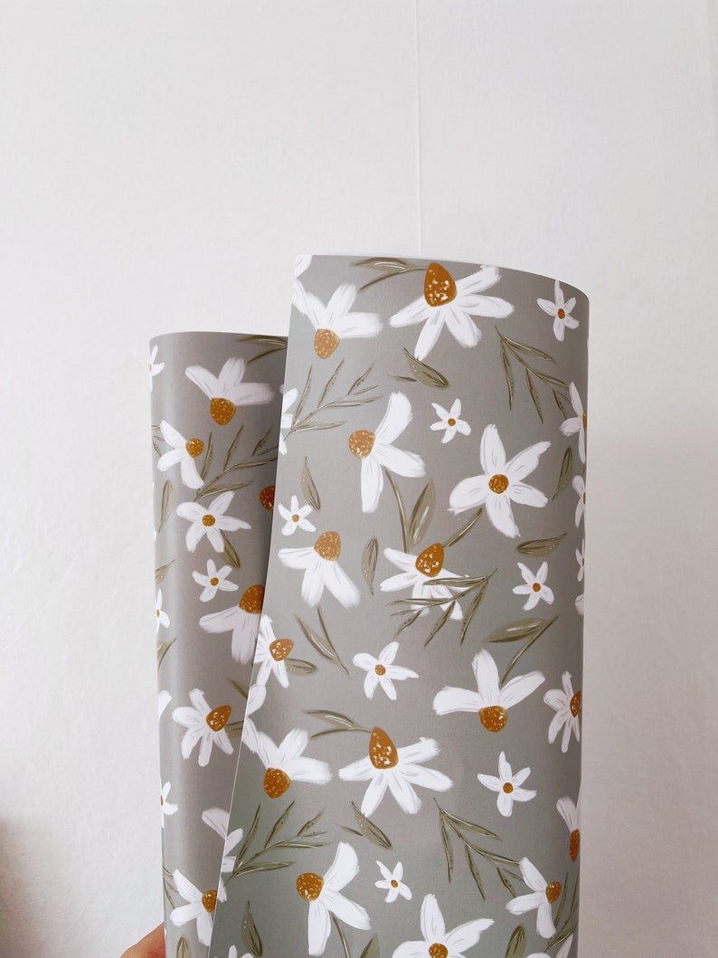 Op de afbeelding: Een rol grijs inpakpapier met een wit madeliefje bloemenpatroon. De madeliefjes hebben gele centra en groene bladeren.