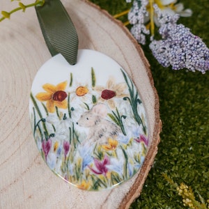 Peut inclure: Une décoration ovale en céramique blanche avec une scène peinte d'une souris dans un champ de fleurs de printemps. Les fleurs sont jaunes, blanches, roses et violettes. La décoration est munie d'un ruban vert attaché au sommet.