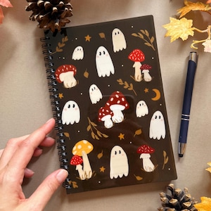 May include: A black spiral-bound notebook with a pattern of white ghosts and red and yellow mushrooms. The notebook is adorned with gold stars and leaf details. A blue pen is placed next to the notebook.