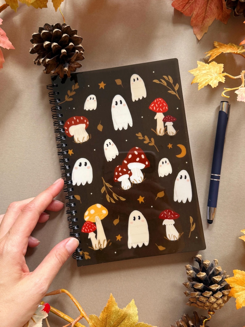 May include: A black spiral-bound notebook with a pattern of white ghosts and red and yellow mushrooms. The notebook is adorned with gold stars and leaf details. A blue pen is placed next to the notebook.