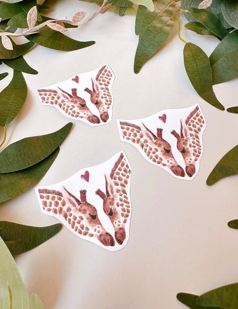 May include: Three watercolor illustrations of two giraffes kissing with a small red heart above them. The giraffes are facing each other and their necks are intertwined. The illustrations are on a white background.