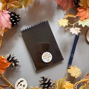 May include: A brown spiral-bound notebook with a sticker reading "Just Smile Designs" is surrounded by autumn leaves, pine cones, and a blue pen. The image has a warm, autumnal feel, with a neutral background.