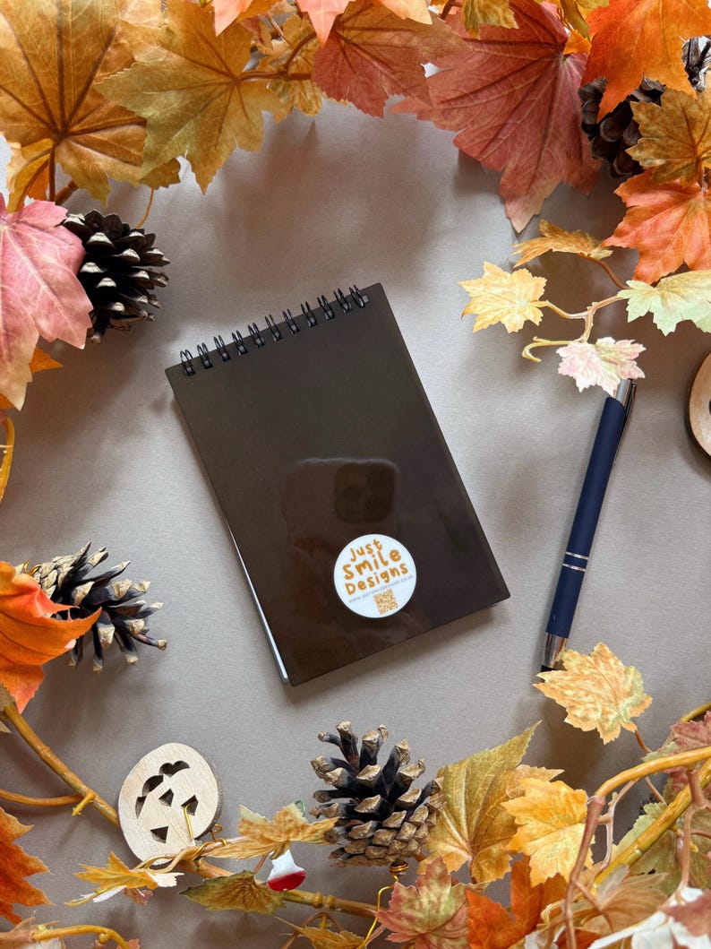 May include: A brown spiral-bound notebook with a sticker reading "Just Smile Designs" is surrounded by autumn leaves, pine cones, and a blue pen. The image has a warm, autumnal feel, with a neutral background.