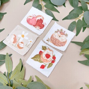 Puede incluir: Cuatro cuadrados de tela blanca con im&aacute;genes bordadas de erizos. Un cuadrado presenta un erizo con una corona de flores, otro tiene un erizo con fresas, otro tiene dos erizos acurrucados y el &uacute;ltimo cuadrado tiene un erizo con margaritas.