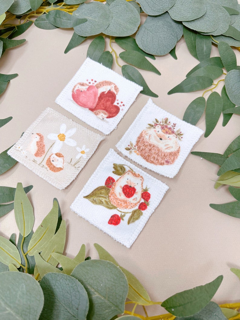 Puede incluir: Cuatro cuadrados de tela blanca con im&aacute;genes bordadas de erizos. Un cuadrado presenta un erizo con una corona de flores, otro tiene un erizo con fresas, otro tiene dos erizos acurrucados y el &uacute;ltimo cuadrado tiene un erizo con margaritas.