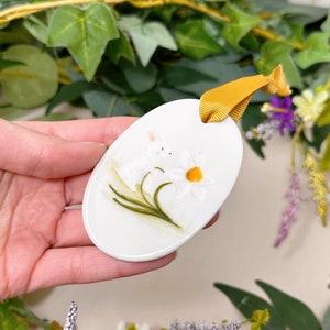 Puede incluir: Adorno ovalado de cerámica blanca con un diseño pintado a mano de dos conejos blancos y un narciso amarillo.