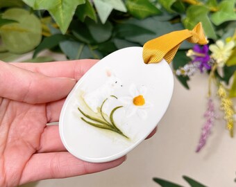 Pode incluir: Ornamento oval de cerâmica branca com um design pintado à mão de dois coelhos brancos e um narciso amarelo.