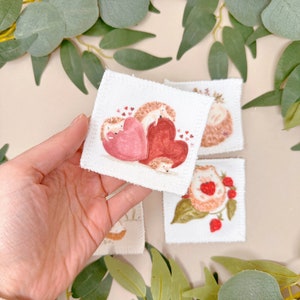 Puede incluir: Cuatro cuadrados de tela blanca con im&aacute;genes bordadas de erizos. Un cuadrado presenta dos erizos sosteniendo corazones rosas y rojos. Otro cuadrado muestra un erizo con fresas. Los dos cuadrados restantes muestran erizos con flores.