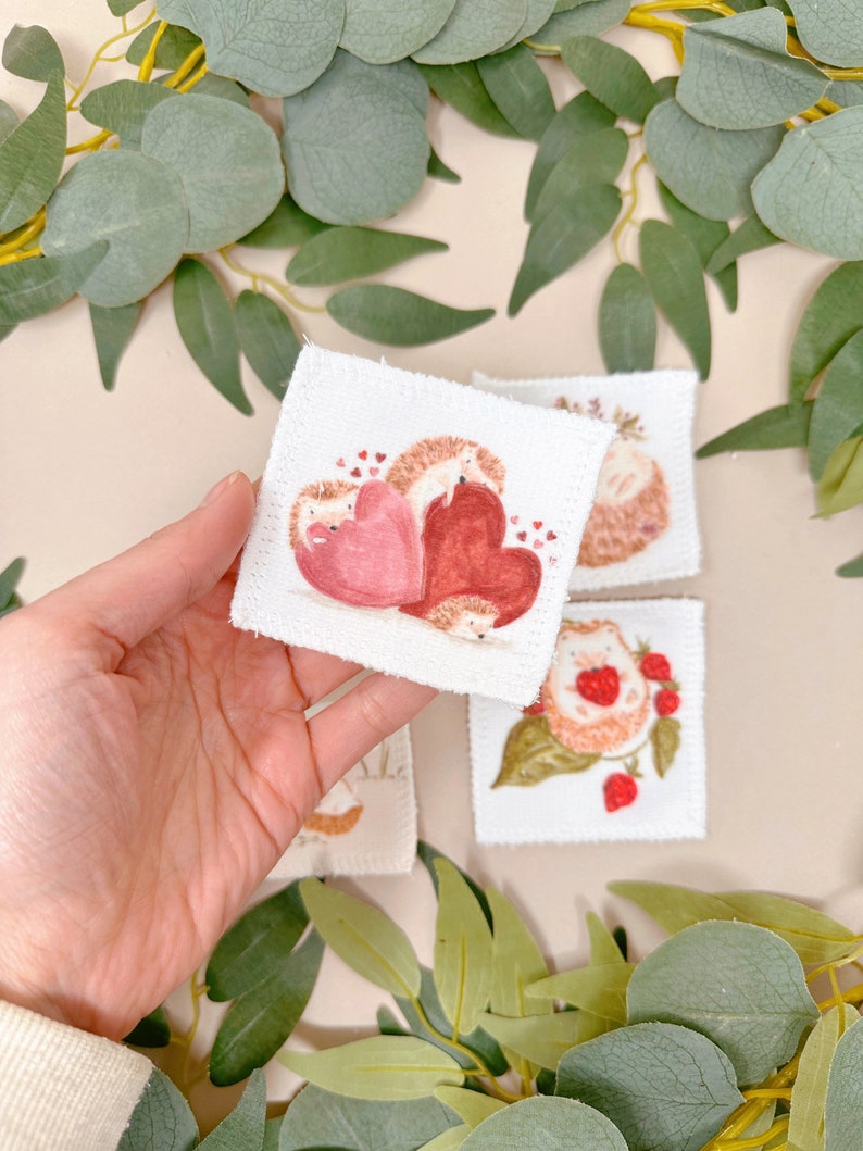 Puede incluir: Cuatro cuadrados de tela blanca con im&aacute;genes bordadas de erizos. Un cuadrado presenta dos erizos sosteniendo corazones rosas y rojos. Otro cuadrado muestra un erizo con fresas. Los dos cuadrados restantes muestran erizos con flores.