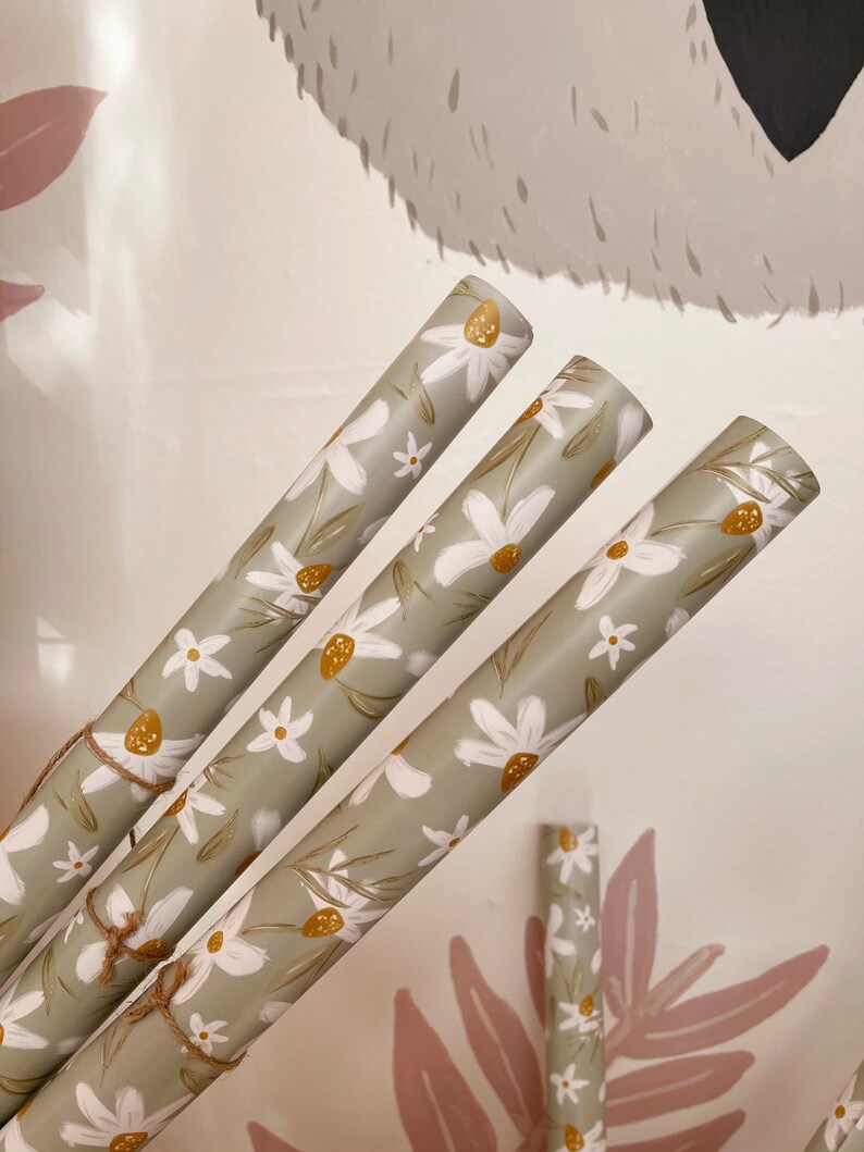 Op de afbeelding: Drie rollen groen inpakpapier met een wit madeliefje bloemenpatroon. Het papier is opgerold en vastgebonden met touw.
