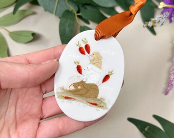 Pode incluir: Ornamento oval de cerâmica branca com um coelho castanho e dois coelhos brancos segurando cenouras. O ornamento tem a palavra "hop" escrita três vezes ao redor da borda.