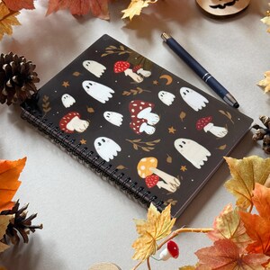 May include: A black spiral-bound notebook with white ghost and mushroom designs. A dark blue pen lies on the notebook. The scene is decorated with autumn leaves, pine cones, and a wooden jack-o'-lantern.