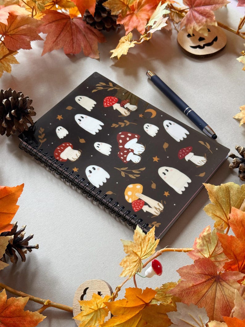 May include: A black spiral-bound notebook with white ghost and mushroom designs. A dark blue pen lies on the notebook. The scene is decorated with autumn leaves, pine cones, and a wooden jack-o'-lantern.