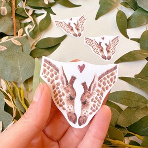 May include: Two watercolor giraffe heads facing each other with a red heart between them. The giraffes are brown and white with a light brown background.