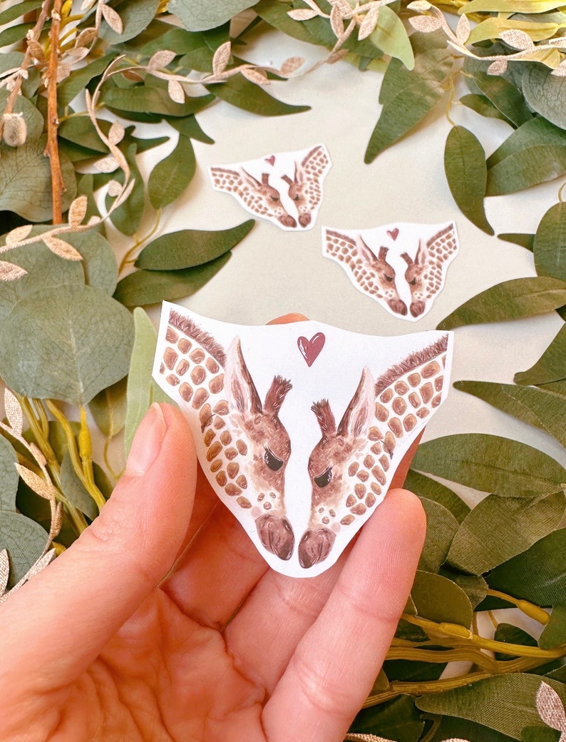 May include: Two watercolor giraffe heads facing each other with a red heart between them. The giraffes are brown and white with a light brown background.