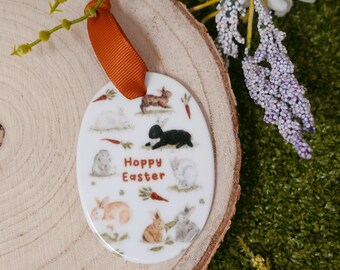 Pode incluir: Ornamento de cerâmica branca com uma fita marrom. O ornamento apresenta um padrão de coelhos e cenouras com o texto "Hoppy Easter" no centro.