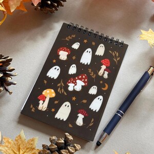May include: A spiral-bound notebook with a dark brown cover featuring illustrations of white ghosts and red and yellow mushrooms. A dark blue pen rests on the right side of the notebook. Autumn leaves and pinecones surround the notebook.