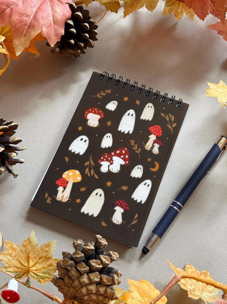 May include: A spiral-bound notebook with a dark brown cover featuring illustrations of white ghosts and red and yellow mushrooms. A dark blue pen rests on the right side of the notebook. Autumn leaves and pinecones surround the notebook.