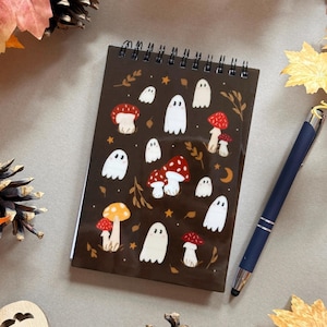 Puede incluir: Un bloc de notas con espiral con una cubierta marrón con ilustraciones de fantasmas blancos y setas rojas y naranjas. Un bolígrafo azul oscuro con punta plateada descansa junto al bloc de notas. El fondo incluye hojas de otoño y piñas.