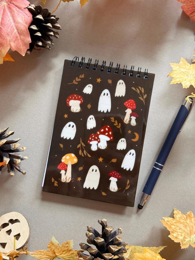 May include: A spiral-bound notepad with a brown cover featuring illustrations of white ghosts and red and orange mushrooms. A dark blue pen with a silver tip rests beside the notepad. The background includes autumn leaves and pinecones.