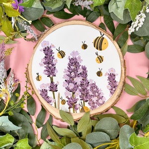 Może przedstawiać: Okrągły drewniany krążek z białym tłem, na którym znajduje się obraz przedstawiający fioletowe kwiaty lawendy i żółte pszczoły. Drewniany krążek jest otoczony zielonymi liśćmi.