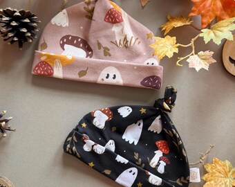 May include: Two baby hats with a fall theme. One hat is light pink with mushroom and leaf patterns. The other is black with white ghost and mushroom patterns. Both hats have a knotted top.