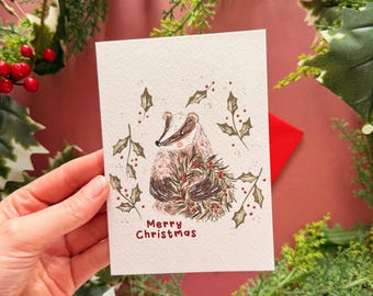 Pode incluir: Um cartão de Natal com uma ilustração de texugo rodeado por azevinho e pinheiro. O cartão tem a inscrição "Merry Christmas" em vermelho. Uma mão segura o cartão, com um envelope vermelho visível atrás. Decoração festiva.