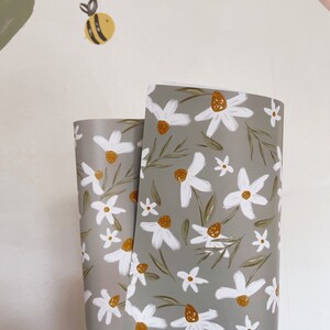 Op de afbeelding: Twee rollen grijs inpakpapier met een wit madeliefje bloemenpatroon. Het papier heeft een subtiel groen en geel kleurenschema.